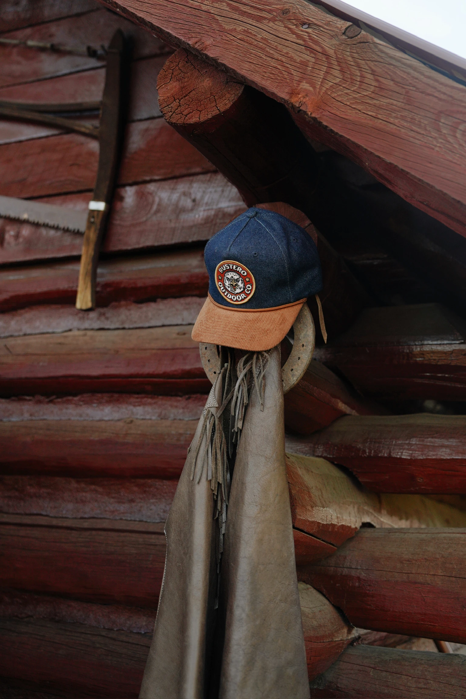 Denim and tan Rustero cap with an embroidered patch, hanging beside a horseshoe against a weathered cabin