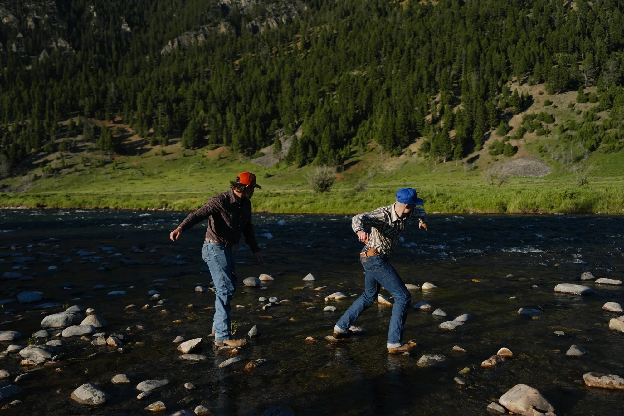 Two people wearing Rustero hats crossing a rocky river, with forest and mountains behind them