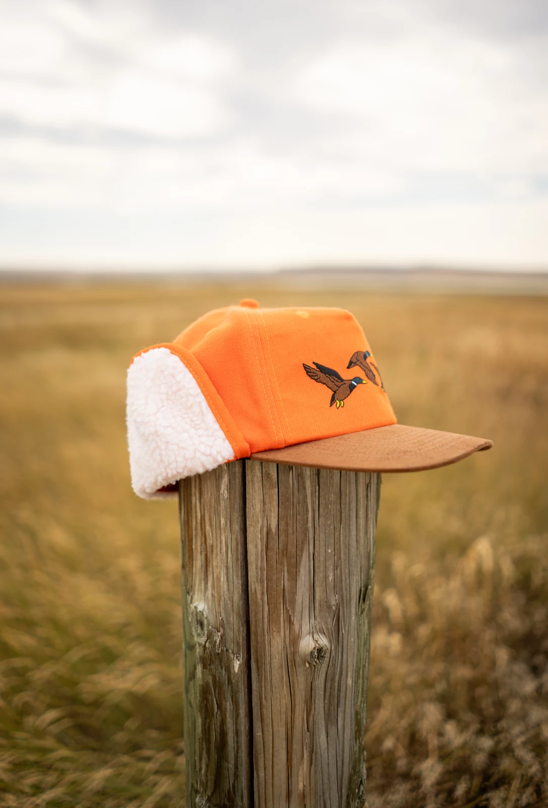 Orange and brown Seager flapjack with embroidered ducks and fleece earflaps, resting on a fence post