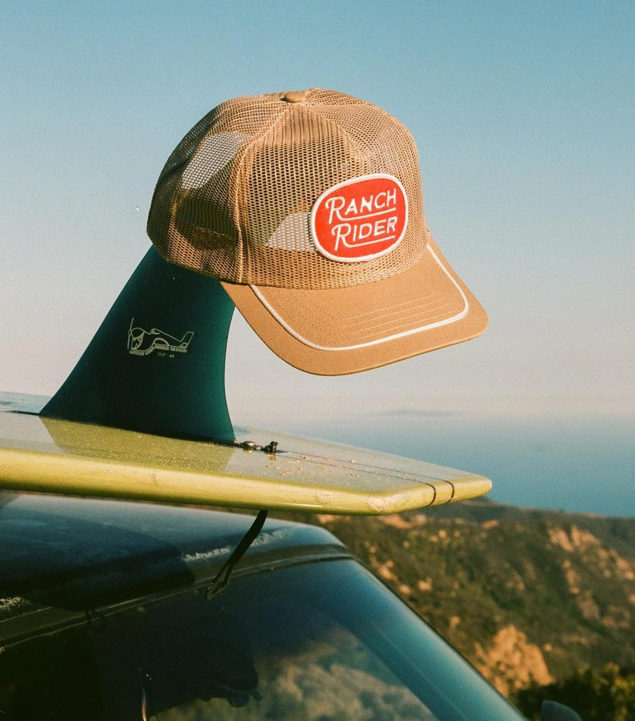 Ranch Rider mesh hat resting on a surfboard fin beside the coast